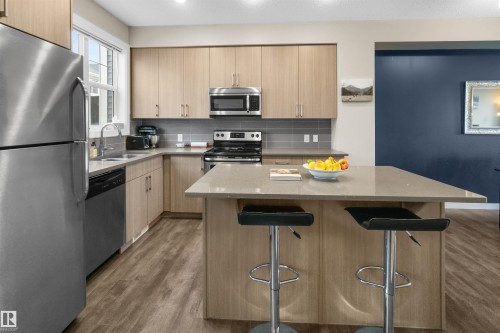 Modern kitchen featuring stainless steel appliances, light wood-finish cabinetry, a large center island with a stone-finish countertop, and wood-finish flooring - 13 2803 James Mowatt Trail, Edmonton, AB 