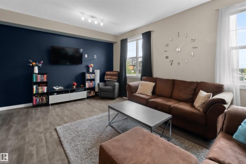 Living area featuring wood-finish flooring, a feature wall with a television mount, and two windows with dark and sheer curtain options - 13 2803 James Mowatt Trail, Edmonton, AB 