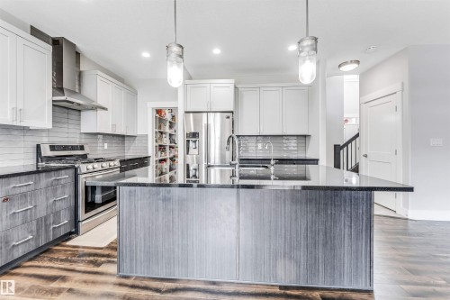 Contemporary kitchen featuring a large island with a dark countertop, white upper cabinetry, grey lower cabinetry, stainless steel appliances, and wood-finish flooring - 8516 216 Street, Edmonton, AB 