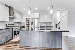 Contemporary kitchen featuring a large island with a dark countertop, white upper cabinetry, grey lower cabinetry, stainless steel appliances, and wood-finish flooring - 