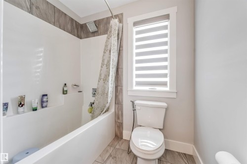 Bathtub and shower combination featuring a tiled surround with a rainfall showerhead, a toilet with a bidet attachment, and a window with horizontal blinds - 8516 216 Street, Edmonton, AB 