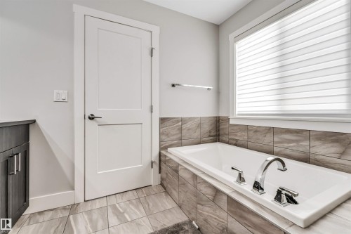 Soaking tub featuring a deck-mounted faucet, surrounding stone-look tile, and a large window with blinds - 8516 216 Street, Edmonton, AB 