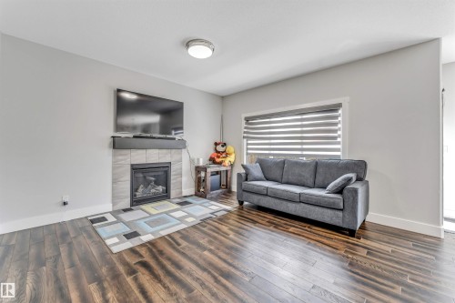 Living area featuring wood-finish flooring, a contemporary fireplace with a tile surround and dark mantel, a large window with horizontal blinds, and a flush-mount ceiling light fixture - 8516 216 Street, Edmonton, AB 