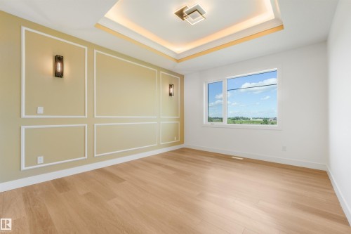Spacious room featuring light wood-finish flooring, a tray ceiling with recessed lighting and a central flush-mount fixture, a paneled accent wall with integrated sconces, and a large window with white trim - 1180 Genesis Lake Boulevard, Stony Plain, AB 