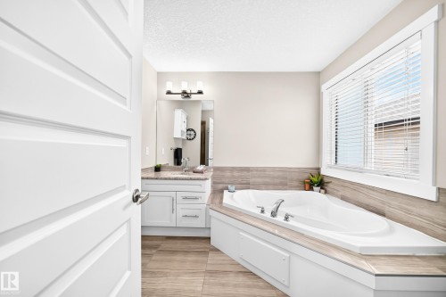 Corner soaking tub with deck-mounted fixtures, framed window with blinds, vanity with undermount sink, stone-finish countertop, and wood-finish flooring - 13724 166 Avenue Nw, Edmonton, AB 