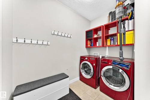 Dedicated laundry area featuring front-loading appliances, wall-mounted red shelving, two wall-mounted coat racks, and light-toned tile flooring - 13724 166 Avenue Nw, Edmonton, AB 
