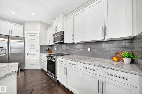 Modern kitchen featuring white cabinetry, light-toned countertops, a full suite of stainless steel appliances, a gray tile backsplash, and dark wood-finish flooring - 13724 166 Avenue Nw, Edmonton, AB 