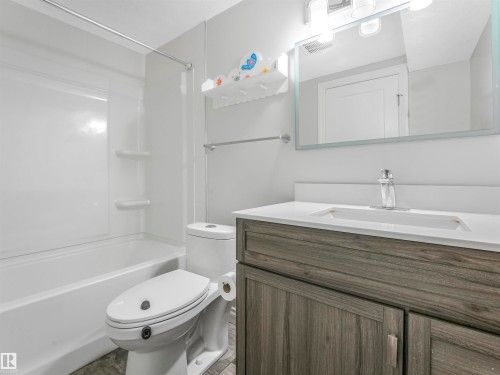 Contemporary bathroom featuring a white tub/shower combination with integrated shelving, a wood-finish vanity with a white countertop and integrated sink, a sleek wall-mounted mirror, and tile-style flooring - 20507 99 Avenue, Edmonton, AB - Indoor Photo Showing Bathroom