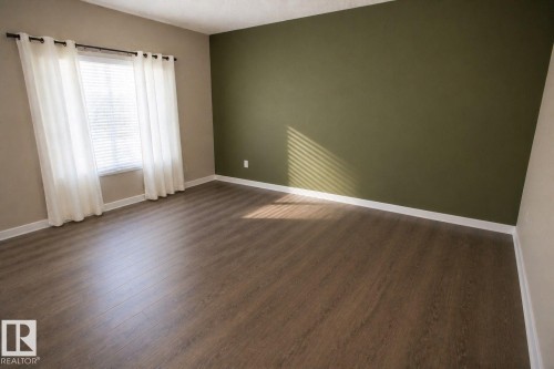 Spacious interior room featuring wood-finish flooring, a window with horizontal blinds, and a contrasting accent wall - 8 Meadowlink Common, Spruce Grove, AB 