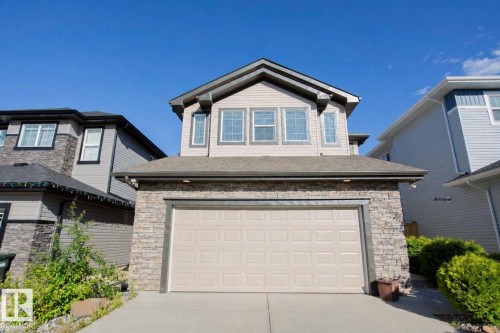 Two-story residential exterior featuring a two-car garage with an overhead door, stacked stone accents, light-toned siding, and multiple upper-level windows - 8 Meadowlink Common, Spruce Grove, AB 