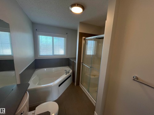 Bathroom featuring a corner soaking tub with a tiled surround, a glass-enclosed shower, a window with blinds, and wood-finish flooring - 8 Meadowlink Common, Spruce Grove, AB - Indoor Photo Showing Bathroom