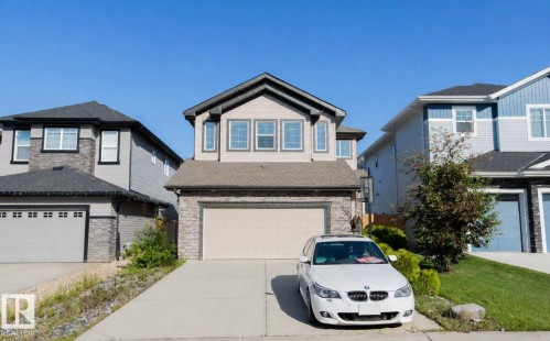 Two-story residence featuring a neutral-toned siding facade with stone veneer accents, a prominent front-facing garage, a concrete driveway, and multiple windows with dark trim - 8 Meadowlink Common, Spruce Grove, AB 