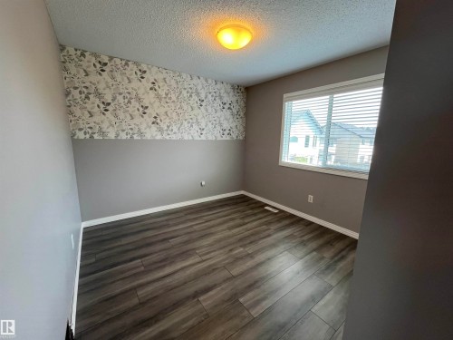 Room featuring wood-finish flooring, a decorative wallpaper accent wall, and a large window with horizontal blinds - 8 Meadowlink Common, Spruce Grove, AB - Indoor Photo Showing Other Room