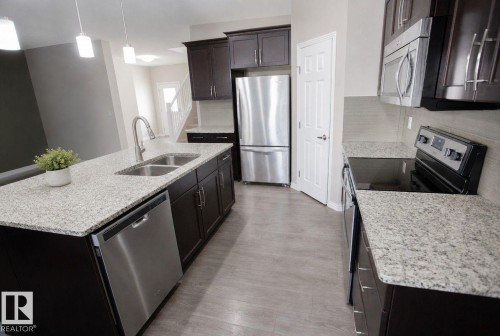 Open-concept kitchen featuring dark wood cabinetry, light speckled countertops, and stainless steel appliances including a refrigerator, dishwasher, and built-in microwave - 8 Meadowlink Common, Spruce Grove, AB 