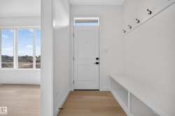 Entryway featuring light wood-finish flooring, a contemporary white door with black hardware, a transom window, built-in bench seating with open storage, and wall-mounted coat hooks - 