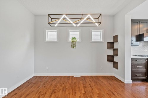 Spacious room with wood-finish flooring, white baseboards, and a contemporary linear chandelier - 614 Mcdonough Link, Edmonton, AB 