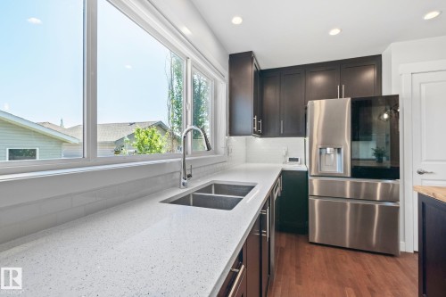 Modern kitchen featuring dark wood cabinetry, stainless steel appliances, a double basin sink with a gooseneck faucet, light-toned countertops, and a subway tile backsplash - 614 Mcdonough Link, Edmonton, AB 