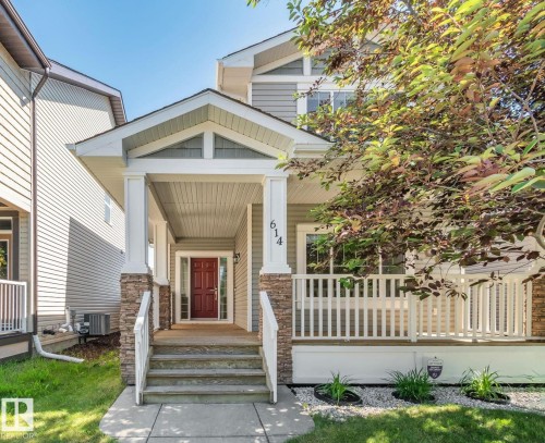 Inviting front exterior featuring a covered porch, stone accents, white railings, a red entry door, and manicured landscaping - 614 Mcdonough Link, Edmonton, AB 
