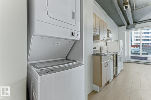 Integrated laundry area with a stacked washer and dryer, complemented by a kitchen featuring light wood-finish cabinetry, white appliances, and a large window - 404 10024 Jasper Avenue, Edmonton, AB 