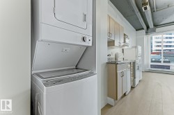 Integrated laundry area with a stacked washer and dryer, complemented by a kitchen featuring light wood-finish cabinetry, white appliances, and a large window - 