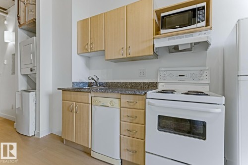Compact kitchen featuring wood-finish cabinetry, granite-style countertops, and a full suite of white appliances including a range, dishwasher, and refrigerator - 404 10024 Jasper Avenue, Edmonton, AB 