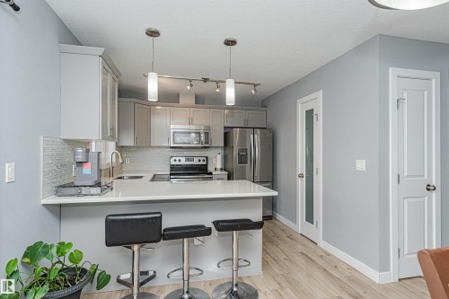 Contemporary kitchen featuring light gray cabinetry, stainless steel appliances, a tile backsplash, light wood-finish flooring, and a breakfast bar with pendant lighting - 35 675 Albany Way, Edmonton, AB 