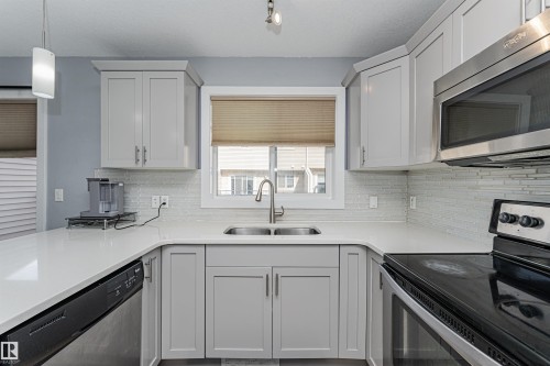 Kitchen featuring light gray cabinetry, white countertops, stainless steel appliances, a tile backsplash, and a double-basin sink - 35 675 Albany Way, Edmonton, AB 