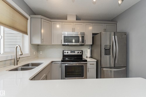 Modern kitchen featuring light gray cabinetry, stainless steel appliances, a double basin sink with a gooseneck faucet, light-toned countertops, and a textured tile backsplash - 35 675 Albany Way, Edmonton, AB 