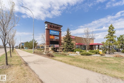 Commercial plaza with a prominent tenant directory sign, established landscaping including mature evergreen and deciduous trees, and a commercial building featuring a red awning - 35 675 Albany Way, Edmonton, AB 