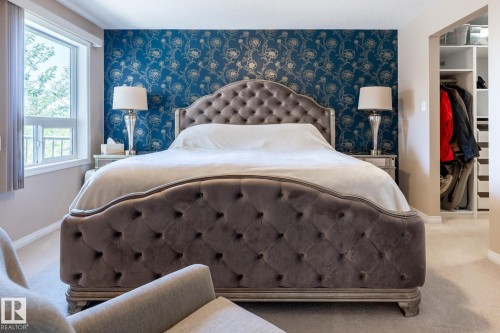 Bedroom featuring a deep blue accent wall with silver floral pattern, light beige wall paint, built-in closet shelving, and a bright window with exterior greenery - 330 1520 Hammond Gate Gate, Edmonton, AB 