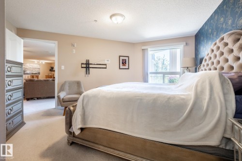 Carpeted room featuring a large window, a partial view of an adjacent room, a wall-mounted light fixture, and a decorative accent wall - 330 1520 Hammond Gate Gate, Edmonton, AB 