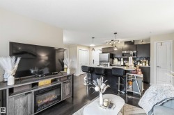 Open-concept living area featuring dark wood-finish flooring, a built-in media console with an electric fireplace, a kitchen island with seating, dark cabinetry, and stainless steel appliances - 