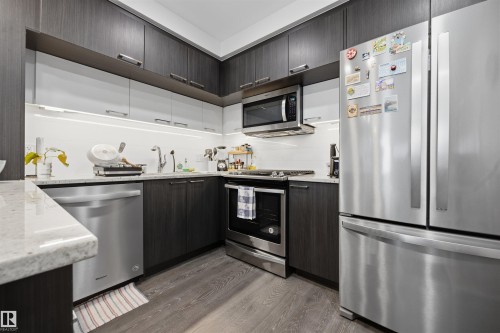 Modern kitchen featuring two-tone cabinetry, light-colored countertops, stainless steel appliances including a refrigerator, dishwasher, and microwave, and wood-finish flooring - 802 9720 106 Street, Edmonton, AB 