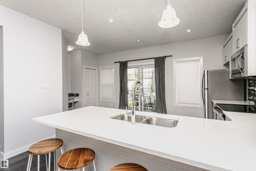Kitchen featuring an expansive white countertop island with an undermount double basin sink, stainless steel appliances, white cabinetry, and a dark tile backsplash - #55 723 172 St Sw, Edmonton, AB - Indoor Photo Showing Kitchen With Double Sink