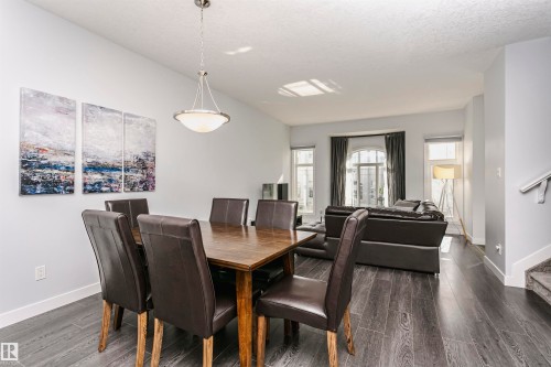 Open-concept dining and living area featuring dark wood-finish flooring, a contemporary pendant light fixture, multiple windows with dark drapery, and a visible staircase with carpeted treads - #55 723 172 St Sw, Edmonton, AB - Indoor Photo Showing Dining Room
