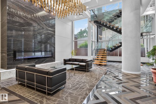 Double-height lobby featuring a modern linear fireplace set against a polished stone accent wall, an open-tread staircase with glass railings, large format windows, and patterned tile flooring - 1504 9720 106 Street, Edmonton, AB 
