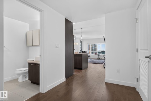 Interior space featuring wood-finish flooring, a white paneled door with louvered details, and a partial view into a bathroom with a tile floor, toilet, vanity with dark cabinetry, and wall-mounted storage - 1504 9720 106 Street, Edmonton, AB 