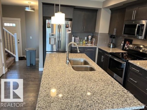 Kitchen featuring a large island with a double basin sink, stone countertops, dark wood-finish cabinetry, and stainless steel appliances - 138 Reed Place, Leduc, AB 
