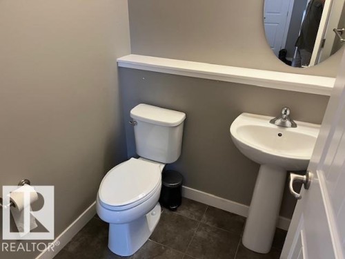 Powder room featuring a white pedestal sink with chrome faucet, a dual-flush toilet, and a circular wall mirror above a white trim shelf - 138 Reed Place, Leduc, AB 
