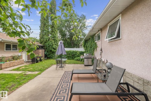 Expansive concrete patio area with a textured stucco exterior, mature privacy landscaping, and multiple awning windows - 12115 53 Street, Edmonton, AB 