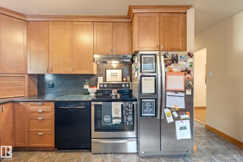 Kitchen featuring wood cabinetry, dark countertops, built-in dishwasher, stainless steel refrigerator, and stainless steel range with overhead ventilation - 12115 53 Street, Edmonton, AB 