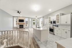 Open-concept living space featuring a stone-clad fireplace with a wood mantel, a central kitchen island with an undermount sink, white cabinetry with dark hardware, stainless steel appliances, and a mix of wood-finish and marble-patterned tile flooring - 
