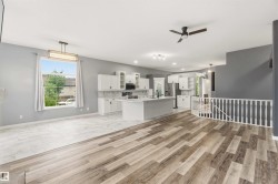 Open-concept living area featuring wood-finish flooring, a contemporary kitchen, white cabinetry, a kitchen island with a sink, and a staircase with white balusters - 