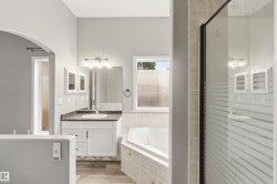 Bathroom featuring a built-in soaking tub with tiled surround, a glass-enclosed shower with frosted detailing, a single vanity with dark countertop and white cabinetry, a large mirror, and wood-finish flooring - 