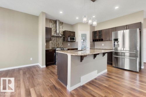 Spacious kitchen featuring a large island with a stone countertop, dark wood cabinetry, stainless steel appliances, a decorative tile backsplash, and wood-finish flooring - 1306 32A Street, Edmonton, AB 