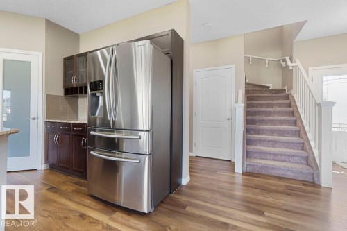 Stainless steel refrigerator with built-in ice and water dispenser, dark wood-finish cabinetry, granite-style countertops, wood-finish flooring, and carpeted staircase with white railings - 1306 32A Street, Edmonton, AB 