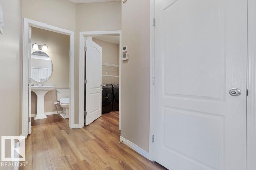 Wood-finish flooring throughout a neutral-toned hallway, featuring white paneled doors, a pedestal sink with an oval mirror, and an in-unit laundry area - 1306 32A Street, Edmonton, AB 