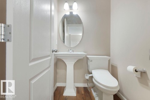Pedestal sink with an oval mirror and wall-mounted vanity lighting, complemented by wood-finish flooring and a white paneled door - 1306 32A Street, Edmonton, AB 