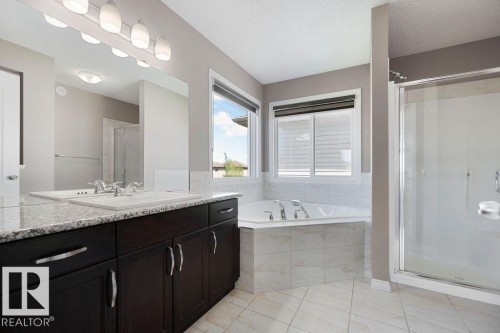 Double vanity with granite countertop and integrated sinks, dark wood cabinetry, corner soaking tub with tiled surround, separate glass-enclosed shower, and tiled flooring - 1306 32A Street, Edmonton, AB 