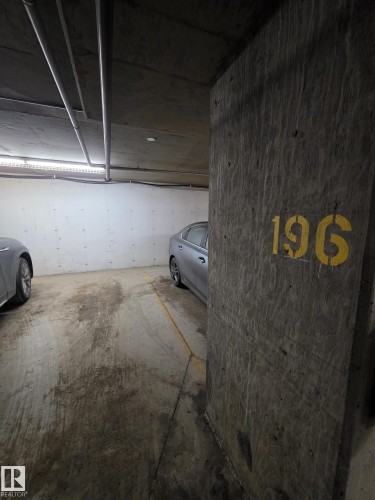 Dedicated parking space within a concrete garage structure, featuring a visible space number, overhead lighting, and exposed utility piping - 310 12045 22 Avenue, Edmonton, AB 